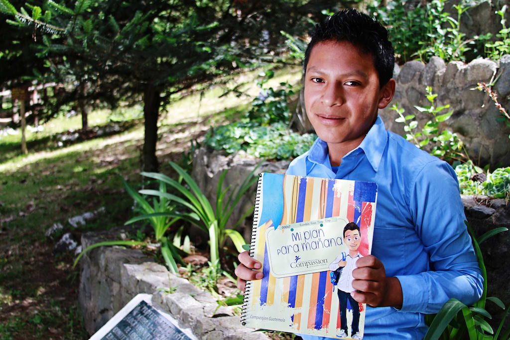 Young boy wearing a light blue button up shirt holds up his workbook sitting beside a garden.